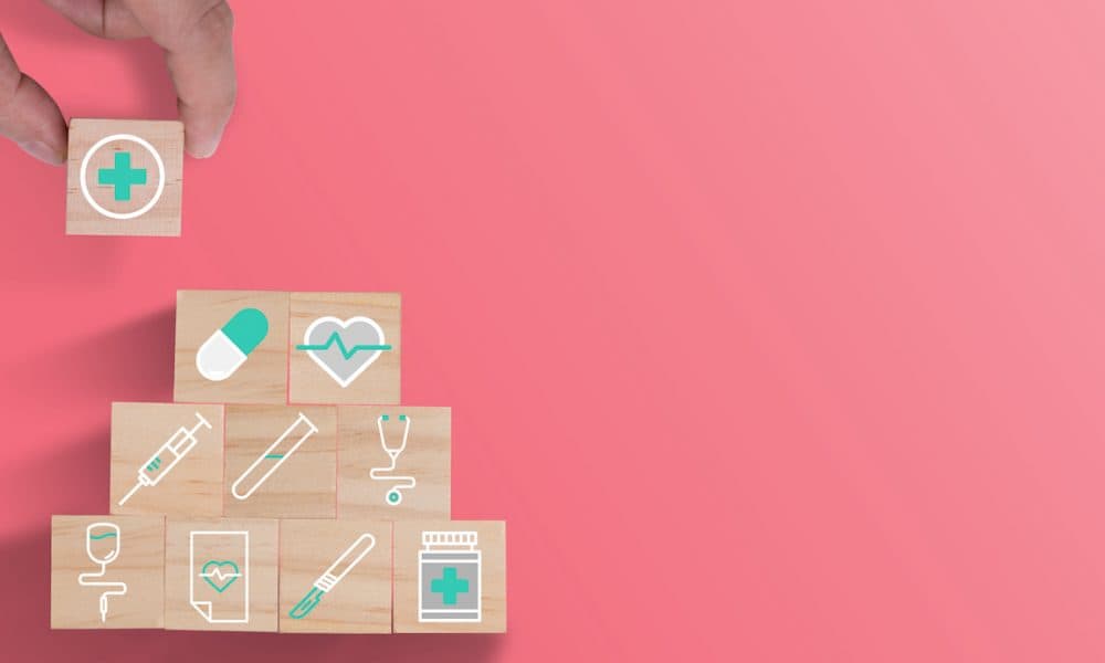Hand putting wooden cubes stacking of healthcare medicine and hospital icon on beautiful pink background. Health care insurance business and investment. New report calls for barriers to be removed in the NHS