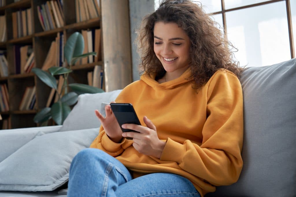 A woman sits scrolling through femtech apps in a yellow jumper and jeans on a couch