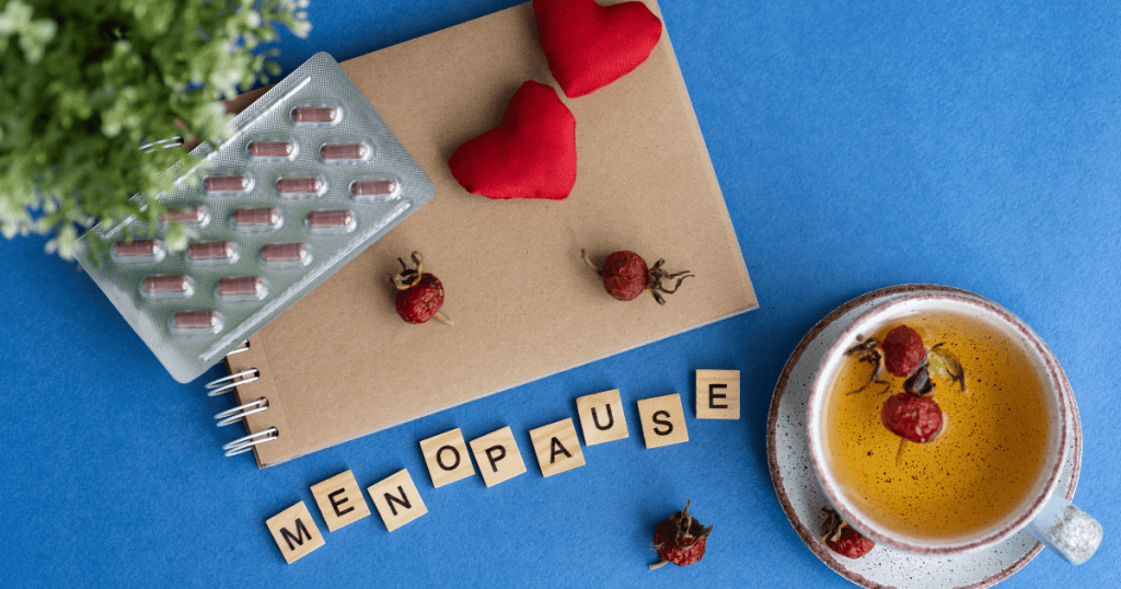 Menopause: A packet of red pills on a notepad next to a cup of herbal tea with red flowers on a blue background. There is a green plant to the left while scrabble tiles spell out menopause
