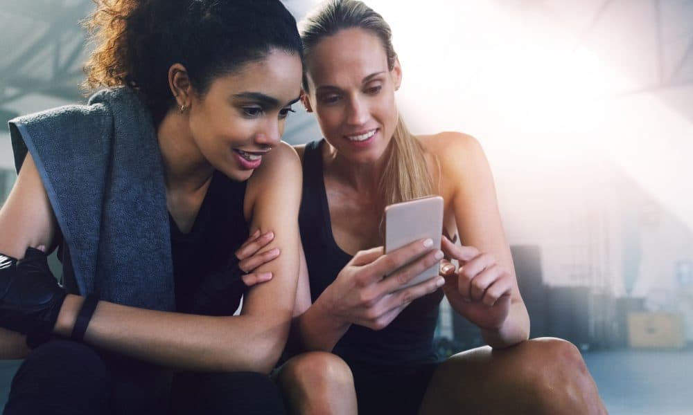 Fitness, phone and girl friends watching a video in a gym with women and smile from training. Social media app, mobile and female person in wellness, exercise and workout class taking a break Financial prescribing: A potential cure to the global inactivity pandemic?