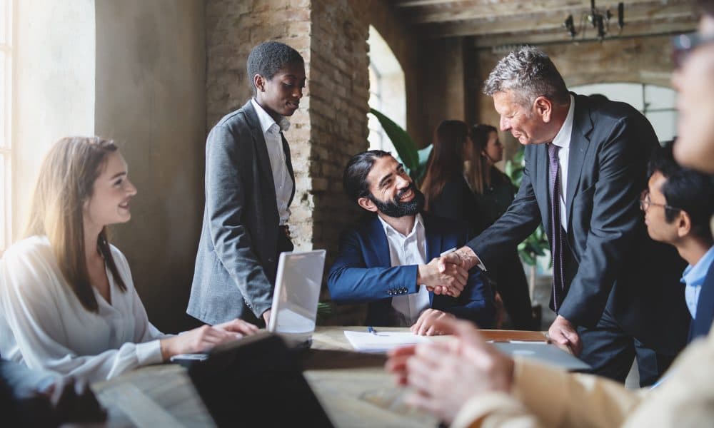 Two businessmen make a deal by shaking hands sitting at a table together with other multiracial employees – business lifestyle and agreements concept Closing healthcare deals in the current market