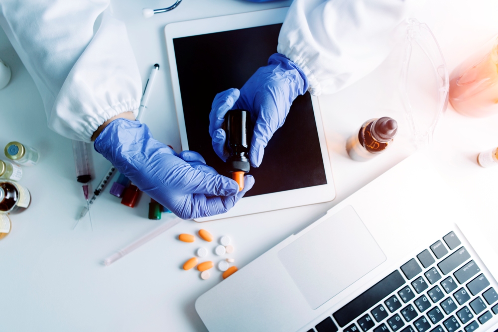 Scientist in blue gloves uses a dropper over a tablet, surrounded by pill bottles and lab equipment on a white workspace.