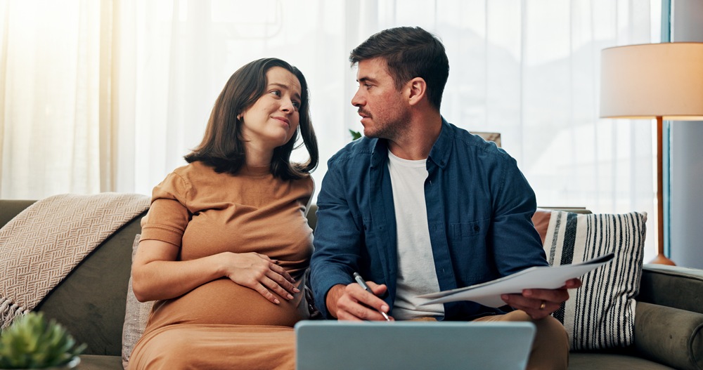 Pregnant woman sits on a couch with her partner; she rests her hands on her belly while he holds papers and a laptop nearby.